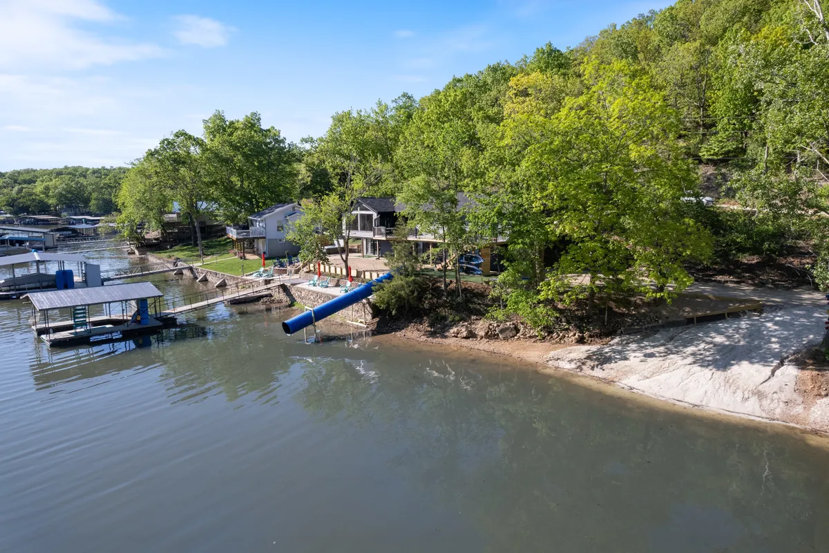 A serene lake with a dock, blue slide, houses, and trees frames a family-friendly setting for boating, fishing, and swimming. The calm water and lush green backdrop evoke a peaceful, playful mood, ideal for both adventure and relaxation.