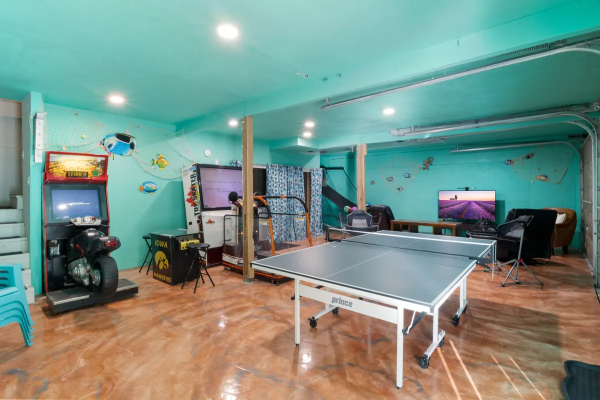 The image showcases an inviting game and arcade room complete with a ping pong table, a classic arcade machine, a TV setup, and some exercise equipment. The space looks spacious and lively, ideal for group activities and all-day entertainment.