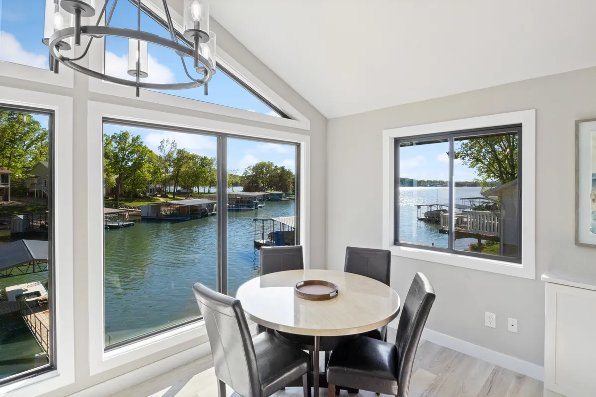 A dining area featuring a large table set beneath a chandelier, surrounded by windows overlooking the lake. Natural light fills the room, and the setting is clearly designed for communal dining with scenic views.