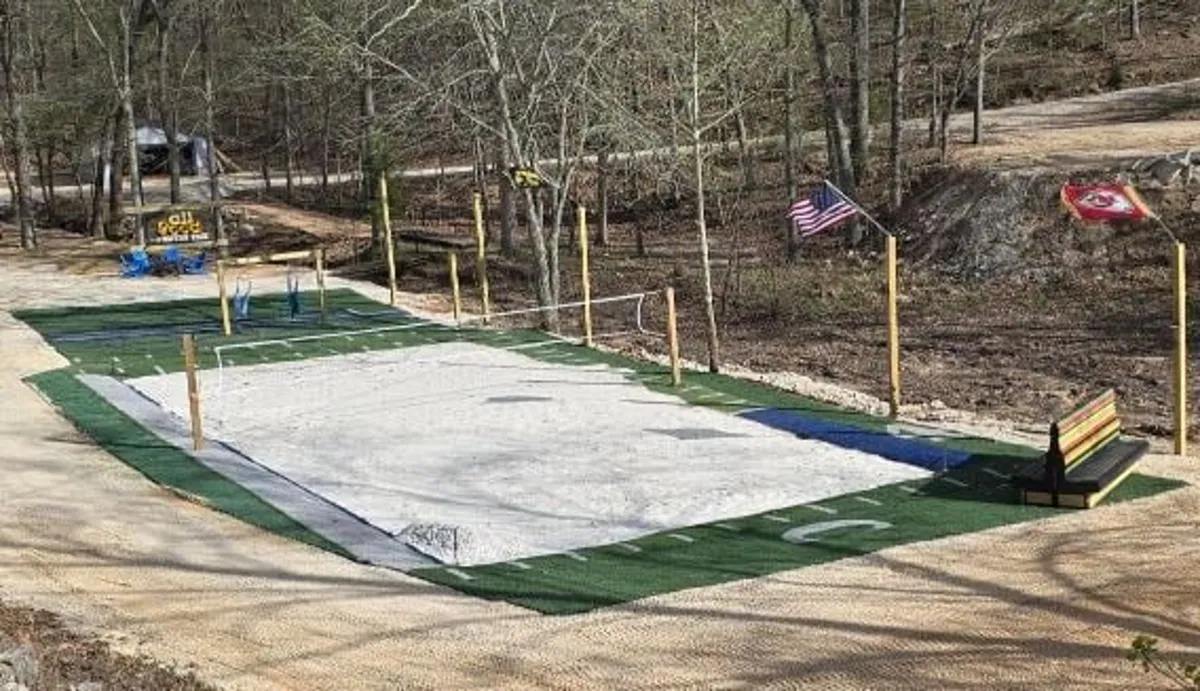 An exterior scene featuring a sand volleyball court surrounded by trees, colorful benches, and playful flags. The lively setup encourages outdoor sports and group games under the sun.