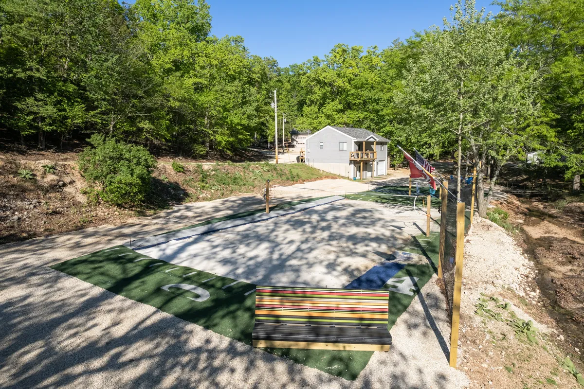 A playful outdoor play area with a clearly marked sports court, a colorful bench, and lush greenery all around. The scene shows ample space for volleyball and other group games, with a small building and trees in the background, making it ideal for family activities and kids’ adventures.