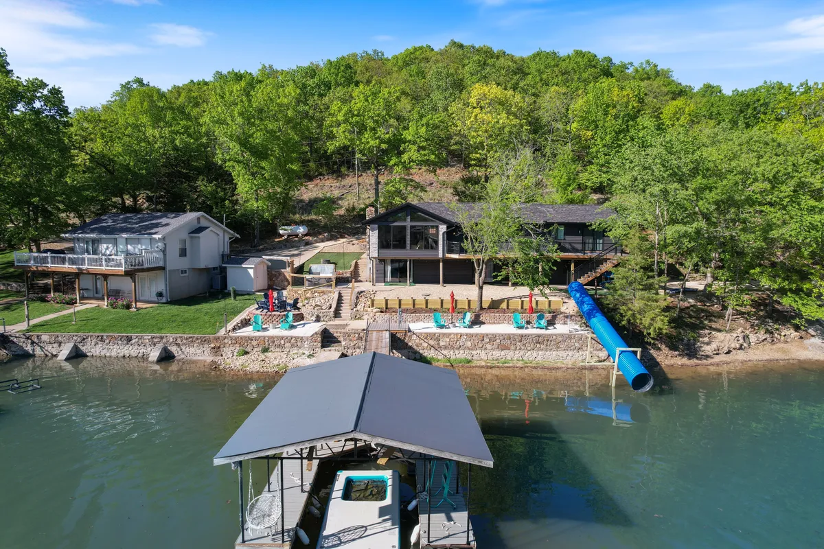 A stunning lakefront property is showcased, with a modern house, a private dock, colorful Adirondack chairs, and a prominent water slide descending into the lake. The scene captures the vibrant waterfront area set up for relaxation and play, with sunset lighting adding to the inviting atmosphere.