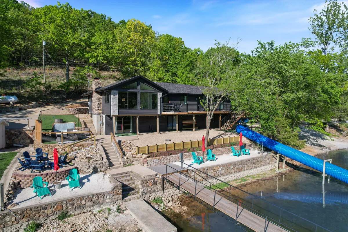 The image showcases a spacious, modern lakefront home with a vibrant blue water slide leading into the lake, an inviting green lawn with comfortable seating, and a private dock stretching out over the water. The sun is setting, casting a warm glow over the entire scene, creating an idyllic atmosphere perfect for outdoor fun and relaxation.