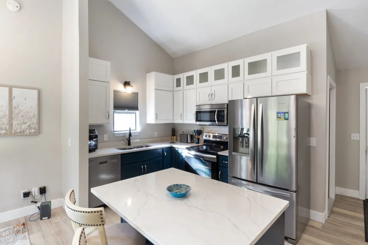 A contemporary kitchen scene featuring a marble countertop, stainless steel appliances, and blue cabinetry. The kitchen feels inviting, with stylish decor and a decorative bowl on the table, evoking a space ready for both meal preparation and casual gathering.
