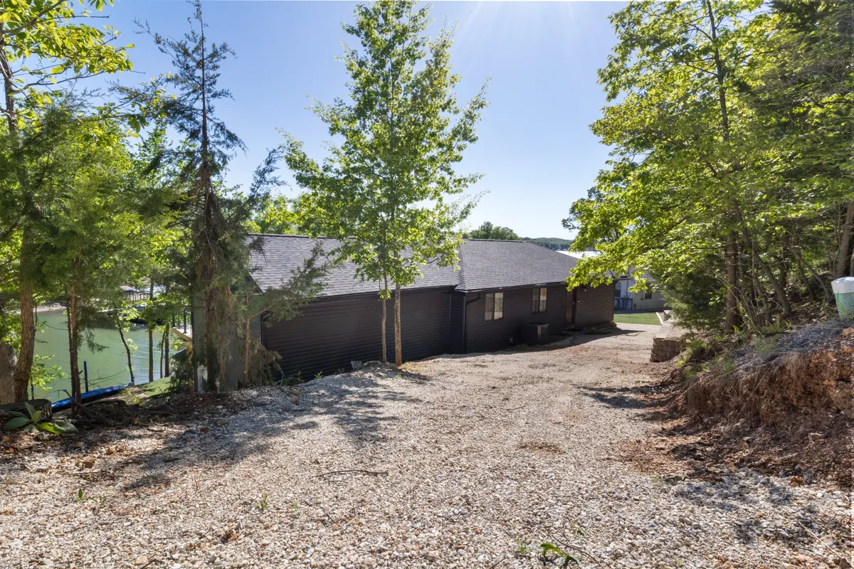 A house facade is nestled beneath a canopy of trees, with a gravel driveway and a clear view toward the lake in the distance. The tranquil scene emphasizes connection to both the home and the natural surroundings.